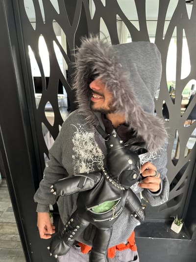 A male individual is depicted in a mid-shot, posing indoors in Marcoussis, France. He faces left, looking slightly upward with a visible smile, and has facial hair. He is wearing a grey hooded sweatshirt featuring a prominent grey fur lining on the hood and white, abstract graphic designs on the left shoulder/chest area. A black, leather-like teddy bear-shaped bag is worn across his front, adorned with numerous silver studs and spikes. This bag also features zippers, one of which is open on the bear's torso, exposing a light green interior lining. The individual's right hand is visible, holding a portion of the bag, and a silver ring is present on his right ring finger. The background consists of a dark, vertically oriented decorative screen with abstract, organic cut-out shapes, through which bright light from an adjacent area or exterior is visible. A small succulent plant in a white, textured pot rests on a dark surface to the lower right of the frame.