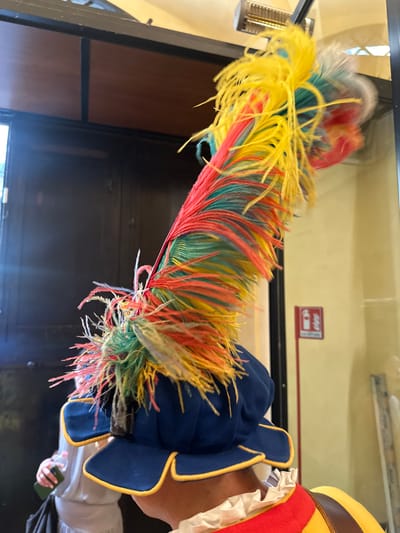 The image features a person wearing traditional Florentine Renaissance clothing, including a blue hat with a yellow trim and a vibrant, multi-colored feather plume. The plume is predominantly yellow, red, and green. The person's collar is visible, appearing white and ruffled. The setting is indoors. There is a dark door or cabinet behind the person, and a fire extinguisher sign is visible on the wall. The overall lighting suggests it is a museum display in Florence, Italy.