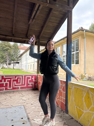 A young woman is captured in a full-body, candid pose outdoors in Troyan, Bulgaria. She stands slightly angled towards the camera, her left arm extended upwards and outwards, and her head tilted slightly. She wears a light blue denim jacket over a black padded vest, dark ribbed leggings, and white sneakers. In her left hand, she holds a black smartphone, its screen displaying multiple small images.
The setting appears to be a courtyard or an outdoor area connected to buildings. To her left, a dark brown wooden pergola structure with slatted wood paneling provides shade. The ground beneath her is a mix of flagstone paving. Immediately to her right, there are distinct painted sections on the ground: a bright yellow area with white geometric lines and a vibrant red area featuring a white maze-like pattern.
In the background, behind the woman, are two buildings. One building features a light yellow facade with multiple windows and a metal gutter pipe running along its roofline. Further back, another building with an off-white or light gray facade and numerous windows is visible. A cloudy, overcast sky dominates the upper portion of the image. Green grass and a tree with sparse foliage are also seen in the distance, indicating an outdoor, possibly institutional or public, environment. The overall mood is casual, set during daylight hours on an overcast day.