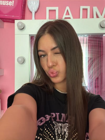 A young woman with long brown hair is taking a selfie in a brightly lit room. She is wearing a black t-shirt and a pearl necklace. Her right eye is closed, and her lips are pursed in a kiss. The background is pink, with white lettering that appears to spell out "ПАПМ". There is a white vanity mirror with lights around it, and a pink box with the word "muse!" on it is visible in the upper left corner. The overall impression is of a well-lit, possibly a beauty or vanity area.