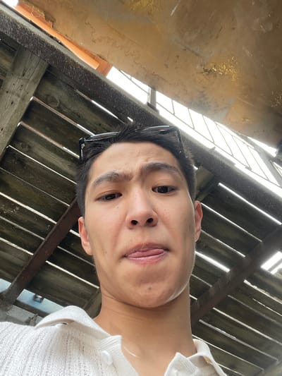 A young person with dark hair and glasses perched on their head is looking up at the camera from an outdoor location. They are wearing a white collared shirt. The background consists of weathered wooden planks forming a slatted ceiling or roof structure, with bright light filtering through the gaps. The image appears to be a selfie taken from a low angle. The location is identified as Кенен Әзірбаев ауылы, Kazakhstan.