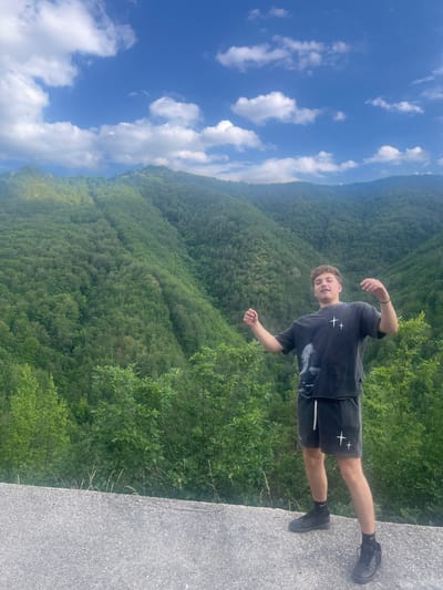 A young man with curly brown hair, wearing a dark grey t-shirt with white crosses and dark shorts, stands on a concrete surface. He is facing the camera with his arms raised slightly. Behind him, lush green mountains covered in trees rise up, with a bright blue sky dotted with white clouds above. The scene is outdoors and appears to be in a natural, mountainous setting.