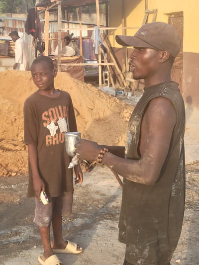 The scene, likely in Kwamba, Nigeria, shows two individuals near a pile of earth or construction material. One individual, possibly a child, wears a brown t-shirt with a "Ghost" graphic, gray shorts, and light-colored sandals. They hold a small white object in their left hand, and an older person, wearing a black sleeveless shirt, a dark baseball cap, and beaded bracelets, is holding a metallic object over them. Dust or particulate matter covers their arms and upper torso. In the background, there appears to be a partially constructed or dilapidated building with corrugated metal sheets. Two other people stand in the background. The overall environment appears to be outdoors with a mix of construction debris and signs of activity.