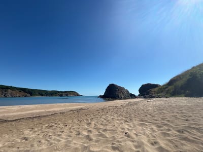 The image depicts a sandy beach in Rezovo, Bulgaria. The foreground is dominated by textured sand with footprints. In the mid-ground, the calm blue sea meets the shore, with several large dark rocks protruding from the water. Further in the distance, a green, tree-covered coastline forms a bay. The sky is a clear, bright blue with a sun flare visible in the upper right quadrant. The overall scene is a serene coastal landscape.