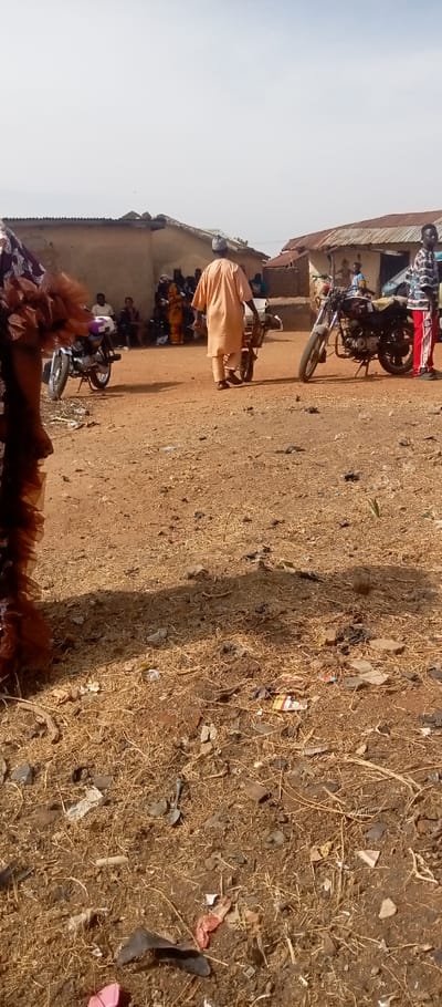 The scene depicts an outdoor area in Kasuwan Magani, Nigeria, under bright daylight with a clear sky. The ground is dry, reddish-brown earth with sparse dry vegetation and scattered debris, including plastic fragments, paper, and small stones.
In the midground, a male figure, viewed from behind, wears a light orange tunic and cap while pushing a two-wheeled hand cart carrying a white container. He moves towards a group of approximately 5-7 individuals seated or standing in the shade of a building on the background-left. One person in this group is visible in yellow attire.
Two motorcycles are parked on kickstands: one light-colored, with a purple and white bag on its rear, is situated in the midground-left; a dark-colored motorcycle is parked in the midground-right. Standing near the dark motorcycle is another male figure wearing a patterned black, white, and red shirt with red striped trousers.
The background features multiple single-story buildings with light-colored walls and corrugated metal roofs. The building on the right exhibits a painted mural depicting figures on its exterior wall. A partially visible person in ruffled brown fabric is present in the far left foreground.