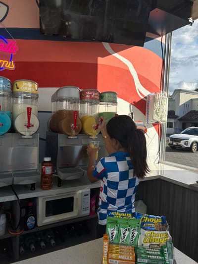 The scene is in a small food stand, likely a street vendor, located in Pozos, Costa Rica (latitude: 9.951, longitude: -84.194). A woman in a blue and white checkered shirt is dispensing a yellow slushie from one of the three machines into a cup in front of her. Two other machines are visible, one with a pink slushie and one with a brown slushie. All the machines are labeled in Spanish. There is a microwave under the slushie machines, along with various bottles and items. There are also packs of Halls and Trident gum on a shelf below the counter. The woman is facing away from the viewer. The stand is open to the street, with a view of a parked car and a building with a brown roof. The stand has a red, white, and blue color scheme and a large flat-screen monitor on the wall behind the woman. The image is taken in the middle of the day, based on the natural lighting and the shadows.
