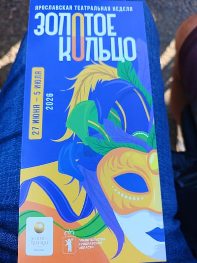 The image shows a close-up of a blue poster held by someone wearing blue jeans. The poster is for the "Yaroslavl Theater Week" and features the title "Zolotoe Koltso" (Golden Ring) in large white letters. There is a graphic of a colorful mask with feathers and leaves. In the lower left corner, there is a white square with a golden circle and the text "Zolotoe Koltso" and "Yaroslavl." To the right of this, there is a shield emblem with a bear and a crown, and text that reads "Pravitelstvo Yaroslavskoy Oblasti" (Government of Yaroslavl Oblast). A yellow strip on the left side of the poster indicates the dates "27 June - 5 July 2026." The poster is displayed outdoors, with visible shadows suggesting a sunny day. The focus is on the poster itself, with the background of jeans and a blurred human leg and arm providing context. The poster appears to be promoting a cultural event in Yaroslavl, Russia.