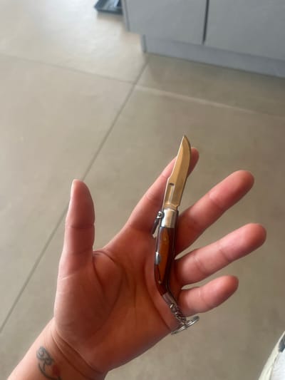 A person's hand is holding a small, wooden-handled pocketknife. The knife is partially open, with its blade extended. A keychain loop is attached to the end of the knife's handle. The hand and knife are resting on a beige, tiled floor. In the background, a light-colored cabinet or counter is visible. The scene is likely indoors, possibly in a residential or commercial setting.