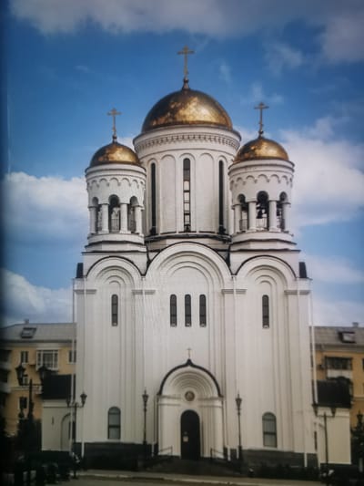 The image shows the Resurrection Cathedral in Serov, Russia. It is a white, multi-domed Orthodox church with golden domes topped with crosses. The cathedral has a central, larger dome and two smaller domes flanking it, each with a bell tower. The architecture features arched windows and entrances, and ornate detailing around the domes and bell towers. In the foreground, there is a paved area with streetlights and some greenery. Behind the cathedral, residential buildings with yellow and gray facades are visible. The sky is blue with scattered white clouds. There are no visible people in the immediate vicinity of the church, though a lone figure is visible in the distance on the left side of the image, near the buildings. The overall scene depicts a prominent religious landmark within an urban environment.