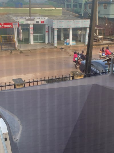 The image depicts a street scene in Shagamu, Nigeria. In the foreground, a grey, textured surface obscures the lower portion of the view. Beyond this, a black metal fence with decorative elements runs horizontally across the frame. To the left of the fence, a concrete pillar with a flat top stands.
In the mid-ground, a street with a wet, muddy surface is visible. Several people are present on motorbikes. On the left, a person in a pink shirt rides a motorbike with two passengers. Further down the street, another motorbike carries at least two people, one in a red top. A dark blue car is parked on the right side of the street.
Behind the street, a building with a prominent sign reading "BARCE GLOBAL NIG LTD" is visible. The building appears to be a shop with glass display windows. Various phone brands like Samsung, Infinix, Tecno, and Itel are advertised on the storefront. Adjacent to this building is another structure with a red sign that includes the word "JECTIONS". The buildings are made of corrugated metal and concrete. In the background, more buildings and vegetation are visible. The sky is overcast.