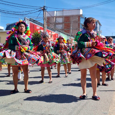 In the city of Wanchaq, Peru, a group of people, primarily women and girls, are participating in a vibrant parade. They are dressed in traditional, brightly colored outfits adorned with intricate floral patterns and layered skirts that flare outwards. Many are wearing elaborate headdresses with colorful tassels and pom-poms. The women are walking on a paved street, casting long shadows in the bright sunlight. In the background, buildings with red and white facades and a large billboard are visible. Power lines crisscross the sky, indicating an urban setting. The overall scene suggests a festive cultural event or celebration.