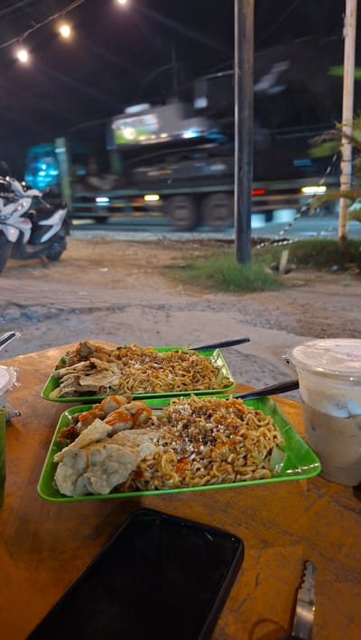 The image shows a dimly lit outdoor scene at night, likely a street food stall or casual dining area. In the foreground, two green rectangular plates of food are set on a wooden table. The plates contain noodles topped with various ingredients, including what appears to be fried dumplings or tempeh, and some are drizzled with sauce. A plastic cup filled with a layered iced beverage is also on the table to the right. In the lower right corner, a black smartphone and a car key are visible on the table.
In the background, a large truck with its lights illuminated is in motion on a road. A white motorcycle is parked to the left of the frame. The environment suggests a roadside setting with dirt or gravel on the ground and sparse vegetation. The overall impression is one of a humble, possibly late-night, dining experience in a public space. The mention of Keudeteupinpunti, Indonesia, places the scene within a specific geographical context.
