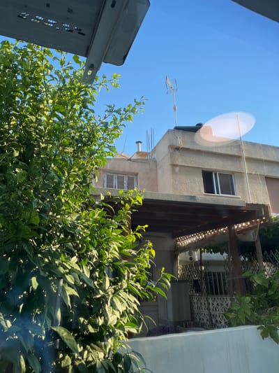An exterior, eye-level, medium shot shows the upper floors of a building in Limassol, Cyprus, partially obscured by lush green foliage from a tree in the foreground. The building's facade is a light, textured plaster, with a second-story window featuring white shutters and a lower-story window with a dark sliding grate. A wooden pergola covers a section of the lower level, supported by columns, and its roof is adorned with decorative latticework. An air conditioning unit is visible beneath the pergola. The rooftop of the building is flat and features a water tank, a television antenna, and several metal rods. The sky is a clear, vibrant blue. A bright lens flare is present in the upper right portion of the image, indicating strong sunlight.