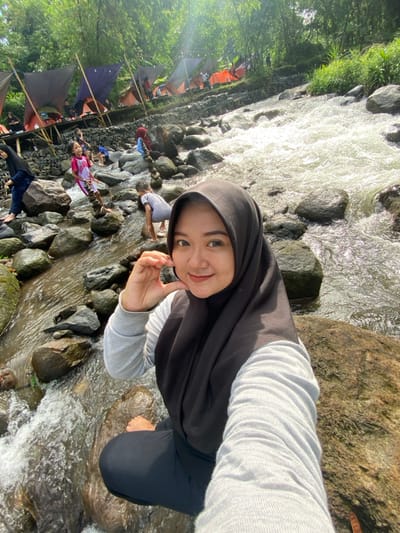 A woman wearing a hijab poses for a selfie while standing in a rocky stream. Clear water flows around her feet and through the rocks. Behind her, a row of tents suggests a campground beside the stream. Other people are visible further down the stream, wading or playing in the water. The scene is bright and sunny, with green foliage visible along the bank.