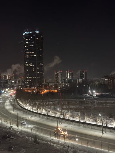 A night cityscape features illuminated buildings including a cylindrical skyscraper dominating the view. Along the right side of the image, a snow-covered road is lined with streetlights and leafless trees. Vehicles traverse the road. The sky is dark.