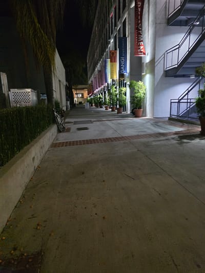 An exterior nighttime shot features a sidewalk next to a building with an outdoor staircase. The sidewalk is paved with concrete and bordered by a low concrete wall. A dense green hedge runs along the wall, and scattered oranges are visible on the ground. To the right of the sidewalk, a building showcases various banners hanging from its exterior, and several potted plants line the walkway. The outdoor staircase, made of metal, ascends along the side of the building, with metal railings and treads. The scene is illuminated by artificial lighting, casting shadows and highlighting the textures of the surfaces.