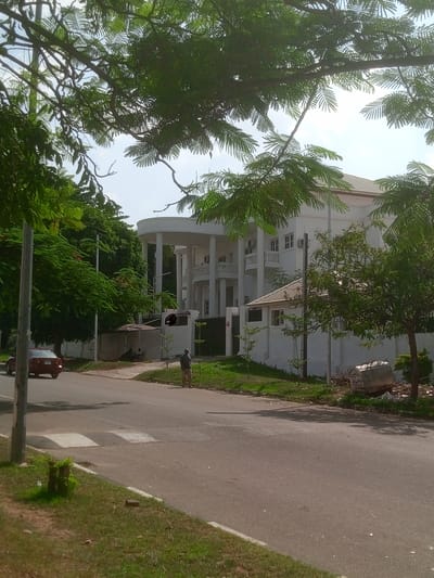 A daytime exterior shot in Aso, Nigeria, depicts a residential street. A large, multi-story white building with columns and balconies is visible behind a white wall and metal fence, partially obscured by trees. A red car is on the left side of the street. A person is walking on the sidewalk in front of the building. Debris is on the side of the street near the building. The street is paved and lined with grass. Trees frame the upper portion of the image.