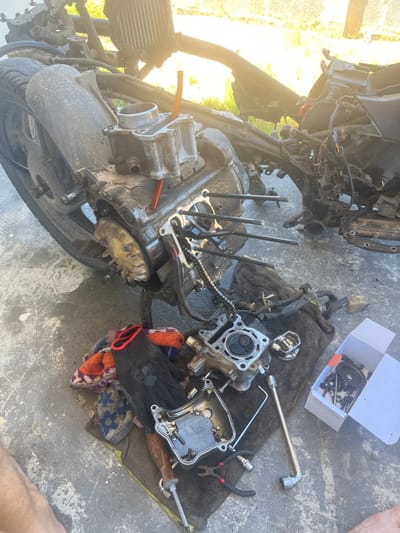 This image captures a motorcycle engine in a state of advanced disassembly on a concrete surface in Cam Đức, Vietnam. The main engine block, heavily soiled, is resting on the ground with its cylinder head removed. Four long cylinder studs protrude from the crankcase, and a timing chain is visible. A detached cylinder block, containing a liner, rests on the engine casing, with an orange plastic tube inserted into it. To the left, a flywheel or stator assembly is partially visible.
Adjacent to the engine, various components and tools are scattered on rags. A cylinder head, displaying its valves and combustion chamber, is positioned on a dark rag alongside a piston and a separate valve cover. Among the tools present are a screwdriver with a wooden handle, a pair of pliers, an Allen wrench, and a socket wrench extension. A multicolored patterned rag is also visible. A white cardboard box, open, contains an assortment of smaller metallic engine parts, including gears and bolts.
The background shows the stripped frame of a scooter or motorcycle, including what appears to be a radiator, with dense green foliage illuminated by bright sunlight beyond. The scene takes place outdoors on a textured gray concrete slab. The lower left corner of the image reveals a bare human foot, indicating the presence of an individual engaged in the repair or maintenance activity.
