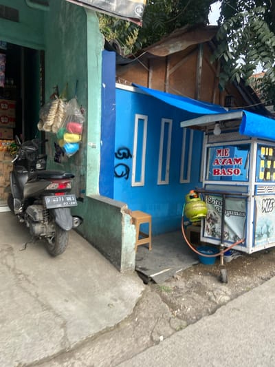 A black scooter with the license plate "D 4271 WEJ 02-26" is parked in front of a green and blue building in Margaasih, Indonesia, at coordinates -6.961, 107.546. The building is painted blue with a green trim. There are three windows on the blue wall and graffiti is visible on the wall below the windows. A yellow propane tank is next to a food cart, which is located on the sidewalk to the right of the scooter. The food cart has a blue tarp roof and says “Mie Ayam Baso" in red letters. There is a wooden stool in front of the blue wall. There is also a small blue plastic basin in front of the food cart. The sidewalk is made of concrete and is cracked in places. The scene is lit by daylight.