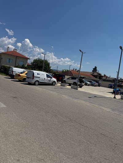 The image shows a street scene in Vedrare, Bulgaria, on a sunny day. In the foreground, a paved road curves to the right. In the middle ground, several vehicles are parked on a paved lot. These include a white van with the logo "ВОЛТРАН" and "www.voltran.bg", a yellow car, a white van, a white Jeep with its trunk open, and other cars. The background features houses with red tiled roofs, trees, and streetlights. The sky is blue with scattered white clouds. A person in a blue shirt is visible in the far right of the image, appearing to be tending to something on the ground.