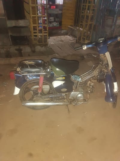 An old, blue and white moped is parked on a dirt ground in front of a store with yellow metal shelves in Wukari, Nigeria. The moped is weathered, with a torn black seat and a red rear shock absorber. The store behind it has shelves stocked with various items, though they are not clearly identifiable due to the low lighting and image quality. The ground is uneven and dusty, suggesting an outdoor, possibly informal, setting.