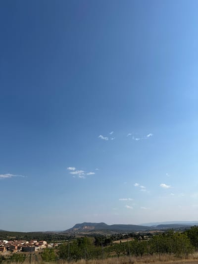 The image depicts a panoramic view of the town of Alpuente, Spain, under a clear blue sky with scattered white clouds. In the foreground, a fence separates the viewer from dry, grassy terrain and the edge of a vineyard or orchard with green trees. Beyond this, the town of Alpuente is visible, characterized by a cluster of buildings with terracotta roofs and stone walls. The town is nestled amidst a landscape of rolling hills covered in green trees and vegetation. In the background, a prominent mountain range rises, with visible geological strata and a distinct plateau shape. The overall scene is one of a rural, sun-drenched environment.