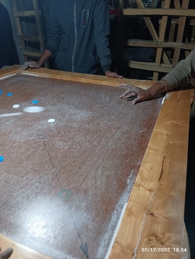 The image displays an indoor scene in Maliat, Bangladesh, centered on a carrom board during play. The board consists of a reddish-brown plywood playing surface, heavily dusted with white carrom powder. Visible carrom pieces include three white circular pieces, three blue circular pieces, and one larger white circular piece, likely a striker.
Two individuals are partially visible. On the left, an individual wearing a dark grey zippered jacket has both hands resting on the light-colored wooden frame of the carrom board. On the right, a second individual, wearing a light brown long-sleeved garment, has their right hand, adorned with a silver bracelet, resting on the board's wooden frame. A third hand is partially visible in the lower-left corner, also resting on the board's frame.
The background is dimly lit and features a dark, multi-tiered wooden shelving or scaffolding structure. The timestamp "02/12/2025 18:54" is visible in the lower right corner of the image.