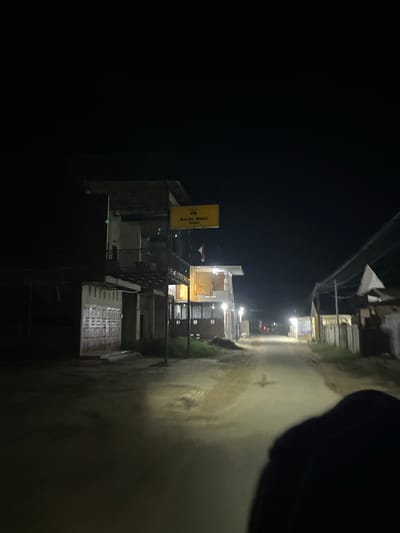The image captures a nighttime street scene in the city of Landuh, Indonesia. A paved road, illuminated by streetlights and possibly vehicle headlights, stretches into the distance, reflecting light on its surface, suggesting it may be wet. On the left side of the road, a two-story building stands, featuring a balcony on the upper level and large metal garage doors on the ground floor. Above the building, a yellow sign with the text "RAJA WALI COFFEE" is visible. A small Indonesian flag is also attached to the building. Further down the street, other structures and utility poles are present, partially obscured by the darkness. In the foreground, the silhouette of a person or an object is prominent, partially obscuring the view of the road. The overall atmosphere is dark and somewhat eerie, typical of a quiet street at night.
