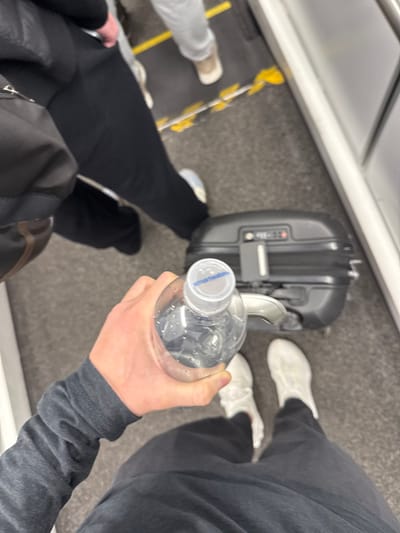 In the city of Boston, United States, a first-person, downward-angle view shows an individual's right hand holding a clear plastic bottle of Smartwater with a grey cap. The individual wears a dark grey long-sleeved shirt and dark pants, with white athletic sneakers on a dark grey textured floor. A black hardshell rolling suitcase with a visible combination lock is positioned directly in front of the individual's feet. To the left, the lower body of a second person is visible, wearing black pants and dark shoes, with a black bag over their left shoulder. Further back and to the right, the lower legs and feet of a third person are seen, wearing light grey pants and light brown shoes. The dark grey floor features yellow safety lines or markings. A grey metallic structural element, likely an interior wall or door frame, runs vertically along the right side of the image. The scene suggests a public transportation environment, such as an airport terminal or boarding area.