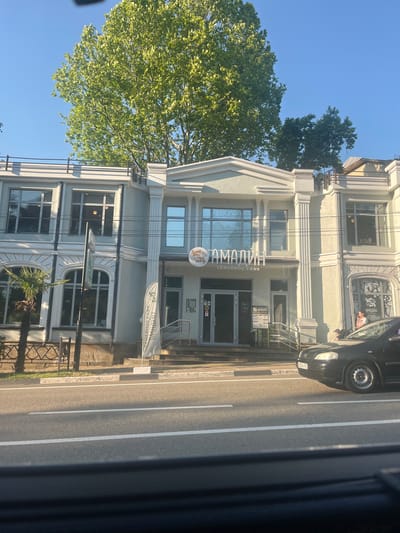 The image captures a street view in Bolshoy Sochi, Russia, with a light-colored, multi-story building prominently featured. The building has a classical architectural style, with columns and decorative elements. A sign above the entrance reads "АМАДИН" and below it "СЕМЕЙНОЕ КАФЕ," indicating a family cafe. A white flag with text is positioned near the entrance. Large windows adorn the facade, reflecting the surrounding environment.

In front of the building, a black car is parked on the right side of the street, and a person is visible near it. A street with white lane markings stretches across the foreground. Tall, leafy trees are visible behind and to the left of the building, casting shadows. The sky is clear and blue, suggesting a sunny day. A palm tree is situated to the left of the building. The overall scene depicts a typical urban setting with commercial establishments and infrastructure.
