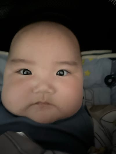 A close-up, head-and-shoulders portrait of a young baby is captured in an indoor setting. The baby's face is the primary focus, filling most of the frame. The baby has round cheeks and a smooth forehead, with dark, alert eyes looking directly at the camera. Their mouth is slightly parted. The baby is wearing a dark blue bib or garment around their neck. In the background, slightly out of focus, are what appear to be baby bedding with a pattern of yellow stars on a gray fabric, and a hint of a light-colored object to the right. The lighting is soft, illuminating the baby's face. The background suggests a domestic environment, possibly a crib or bassinet, within a home in Тайгараев, Kyrgyzstan. No specific actions or interactions are evident, other than the baby's direct gaze.