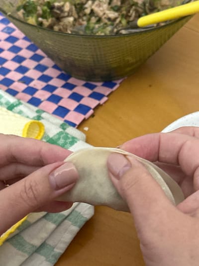 The image presents a close-up, high-angle view of hands meticulously folding a dumpling. A pair of hands, featuring light pink manicured nails, are centered in the frame, delicately shaping a light-colored dumpling, which appears to contain a filling. The hands are positioned above a patterned cloth with a green and white checkered design, which in turn rests on a light brown wooden surface, possibly a table or countertop.

To the left of the hands, another fabric with a distinctive pink and blue diamond pattern is visible. Further left, a dark green or black ribbed object, resembling a bowl or container, is partially seen, with a bright yellow handle of a utensil, likely a spoon or spatula, protruding from it. The far-left background is blurred, showing indistinct outdoor elements like foliage or ground, suggesting a window or outdoor view adjacent to the food preparation area. The scene depicts a domestic setting, likely a kitchen or dining area, focused on the culinary activity of making dumplings. This event takes place in Egkomi Lefkosias, Cyprus. No specific time of day, weather, or visible text can be discerned. The image conveys a sense of focused and calm food preparation.