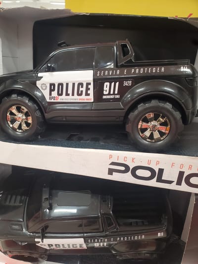The image captures a black toy police truck in its retail packaging, viewed from an angled perspective. The toy car is rugged in appearance, featuring large, knobby black tires with shiny, chrome-like spoked rims. The vehicle's body is predominantly black with white decals.

Visible text on the toy includes the word "POLICE" in large white letters on the side, accompanied by "911" and "EMERGENCY CALL." Below "POLICE," the acronym "RPDSF" and the phrase "ROMA POLICE DEPARTMENT SPECIAL FORCES" are printed, along with a circular emblem. Vertically along the side, the Portuguese phrase "SERVIR E PROTEGER" (meaning "To Serve and Protect") is present, aligning with the given location context of Curitiba, Brazil. The number "3426" is also visible on the side.

The packaging itself is partially visible, featuring large, vertical lettering that appears to be part of the word "POLICE," and above it, "PICK-UP FOR." The box design includes white text on a dark background, possibly black or dark gray, with some red/orange accents. The background behind the box is a solid yellow on the left side of the frame, suggesting the item is displayed in a retail environment. The image is rotated 90 degrees counter-clockwise. No activity is taking place; it's a static product shot.