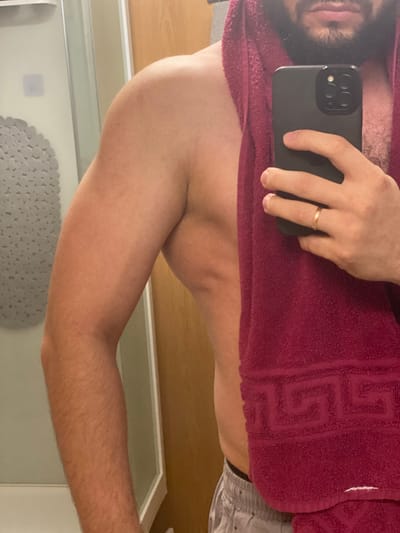 In a close-up, waist-up shot, a person with dark hair and a beard is reflected in a mirror. They are wearing a light-colored towel draped over their shoulder and a gold ring on their left ring finger. The person is holding a black smartphone in their right hand. The background includes a white and grey patterned shower curtain and a wooden doorframe. The scene appears to be indoors, likely a bathroom. There is no discernible indication of the city of Koskolovo, Russia.