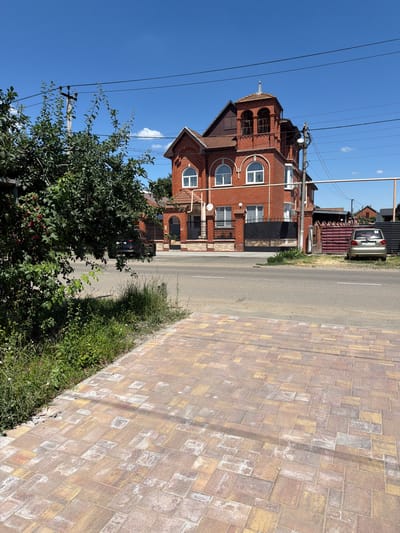 The image depicts a residential street scene in Timashevsk, Russia, under a clear blue sky. In the foreground, a paved walkway made of multi-colored rectangular stones leads towards the right. Adjacent to the walkway is an area of green grass and weeds.
Dominating the middle ground is a large, multi-story brick house with a distinctive tower-like section featuring arched windows. The house has a tiled roof and a black metal gate with an ornate design. A black SUV is parked on the street in front of the house, partially obscured by foliage from a large green tree on the left side of the image. To the right of the house, a lighter-colored sedan is parked on the side of the road.
Electrical poles and overhead wires are visible against the sky. Further down the street, other residential buildings and fences can be seen. The overall impression is of a sunny day in a suburban or semi-rural area.