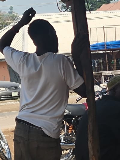 In Suleja, Nigeria, a man wearing a light-colored t-shirt and dark pants stands with his back to the camera, his right arm raised and hand clenched. He appears to be holding onto a wooden pole. Beside him, a motorcycle is visible, with another person partially visible in the foreground wearing a green cap and dark clothing. In the background, a dark-colored car is parked on a dirt road, with buildings and blue tarpaulin structures further beyond. The overall scene suggests an outdoor setting possibly in a market or along a street.