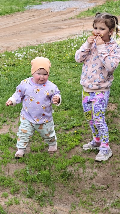 Two young girls are standing on a patch of grass and dirt. The younger girl, on the left, wears a light orange hat, a lavender sweater with rabbit and strawberry designs, and light blue pants with fox designs. She holds a dandelion in her right hand. The older girl, on the right, wears a pink unicorn hoodie and purple, green, and yellow leggings with My Little Pony designs. She eats a piece of food with both hands. The background includes green grass with dandelions and a dirt road. The location is Уральское, Russia.