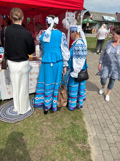 In the city of Podlewkowie, Poland, at a local event, two women dressed in traditional blue and white embroidered folk costumes stand near a red tent. The costumes feature intricate geometric patterns on the sleeves and skirts, with white headpieces adorned with large white bows. One woman wears a black shirt and a long, flowing white skirt, while the other has a white shirt with embroidered blue details. They are positioned in front of a table covered with a white tablecloth featuring floral embroidery. A woven circular mat is visible on the ground near their feet. In the background, other people are browsing stalls, and a building with a gray roof is visible, bearing a sign that appears to be in Polish. The ground is a mix of grass and a paved walkway.