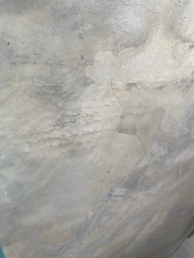 The image shows a close-up, abstract view of a weathered concrete surface. The concrete exhibits a mottled texture with variations in gray and white tones, suggesting age and exposure to the elements. There are numerous small pores and some subtle discoloration, creating a distressed and uneven appearance. A thin, hairline crack is visible in the upper portion of the frame. The lighting is diffused, casting soft shadows that accentuate the textured surface. There are no identifiable objects, people, or actions depicted. The context provided about the city of Tuapse, Russia, cannot be visually confirmed from the image itself, as it solely focuses on the texture and pattern of the concrete.