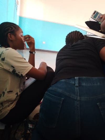 Two people are seated, indoors. The person on the left wears a light-colored t-shirt and black leggings and touches her face with her right hand. A black bracelet is visible on her left wrist. The person on the right wears a black t-shirt and jeans. A calculator sits on a surface to the upper right. The background is a light-colored wall over a teal wall. The room appears to be located in Umueshi, Nigeria.