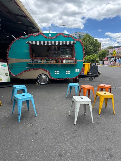 An aqua-colored food truck with a striped awning and red tinsel trim is parked on asphalt. Six metal stools in yellow, orange, white, and blue are arranged nearby. A utility cart and other equipment are behind the truck. Buildings and trees are visible in the background, with a blue sky and clouds above. The image is rotated to the side.