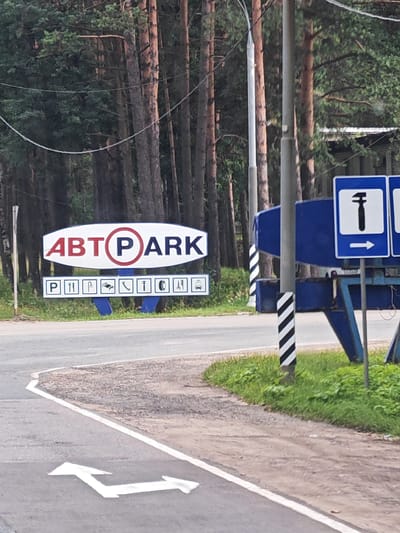 In Yedrovo, Russia, a roadside sign for "ABT PARK" is prominently displayed. The sign features "ABT" in red lettering and "PARK" in blue, with a red circle and blue inner circle around the "P". Below this main sign, a smaller sign lists various icons representing services, including a parking symbol, a tire, a wrench, and other pictograms. To the right of the "ABT PARK" sign, a blue structure is visible, and a blue and white striped pole stands nearby. Further to the right, a blue square sign with a white hammer icon and a white arrow pointing right is mounted. In the foreground, a road with white lane markings and a white arrow indicating a turn is visible. The background consists of a dense forest of pine trees.