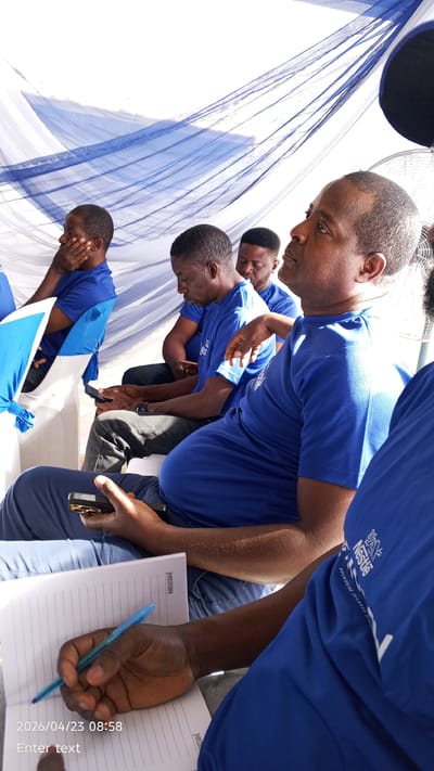 This media file captures a group of men attending an event or meeting, likely in Tarauni, Nigeria. The setting features white and blue draped fabric overhead, suggesting an indoor or tented venue.

The primary subjects are several adult men, all dressed in matching royal blue short-sleeved t-shirts. In the foreground, a man is looking upwards and to the right, appearing attentive or contemplative. His blue t-shirt prominently displays the white Nestlé logo and the partially visible slogan "NOURISHING CHILDREN". Adjacent to him, another man is focused on a mobile phone held in both hands. Further back, other participants are seated, with one resting his hand on his chin, seemingly listening. A white electric fan is partially visible in the upper right, suggesting the need for cooling.

In the bottom left foreground, hands are visible holding a mobile phone and a blue pen, actively writing in an open, lined notebook. The notebook pages are branded with "NESCAFÉ". The activity suggests a session where participants are observing, taking notes, or engaging with their devices.

Visible text includes the date and time "2026/04/23 08:58" in the bottom left corner, "Enter text" (likely a camera placeholder), "NESCAFÉ" on the notebook, and the "Nestlé" logo with "NOURISHING CHILDREN" on a t-shirt. The coordinated attire and branded items point towards a corporate or organized gathering.