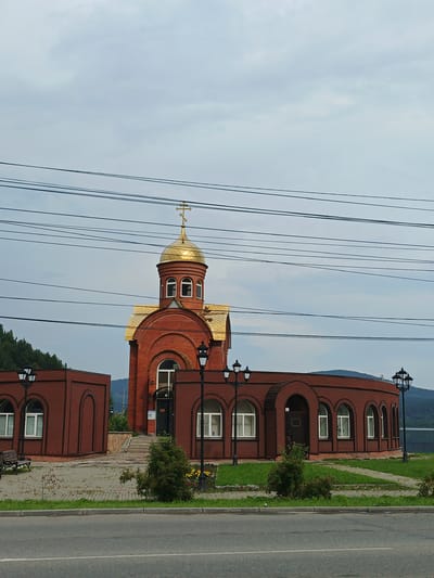 A brick church with a golden dome stands prominently in Zlatoust, Russia. The church features arched windows and a tiled roof. In front of the church, there is a curved brick structure also with arched windows and a dark entrance. The church is surrounded by a paved area with lampposts, benches, and some vegetation. Power lines are visible crisscrossing the sky above the church. In the background, there are trees and a body of water, suggesting a scenic location. The overall scene is captured on an overcast day.