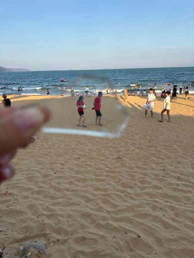 The image captures a vibrant, sun-drenched beach scene at South Quy Nhon Ward, Vietnam, likely during the late afternoon given the warm, golden light and long shadows cast on the sand. A diverse crowd of people are enjoying a day at the beach. Many are in the shallow ocean water, swimming and playing in the gentle waves. Others are on the sandy shore, walking, socializing, or relaxing.
The beach is characterized by its golden sand, which shows ripples from the receding tide and scattered debris like small shells and pebbles. The ocean is a deep blue, with white foam cresting on the incoming waves. The sky above is a clear, pale blue, indicating fair weather.
Several individuals are prominently featured. Near the water's edge, a man in a red shirt and black shorts stands facing away from the camera. Further up the beach, two people in red shirts and dark shorts are walking. A person in a white shirt and shorts is also visible walking on the sand. Further back, a cluster of people are wading in the water, and many more are scattered throughout the scene, indicating a popular recreational spot.
There are no visible signs or text within the image. The overall mood is one of leisurely enjoyment and a typical summer day at a busy beach. The perspective is from someone standing on the sand, looking towards the ocean and the crowded shoreline. A blurry foreground suggests the presence of the photographer's hand or an object in close proximity to the lens.