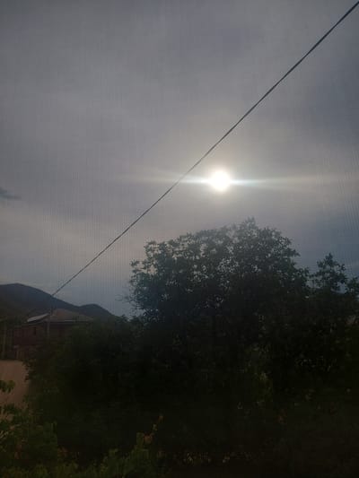 The image shows a view from a window with a screen, looking out at a landscape. The sun is visible in the sky, partially obscured by clouds and a power line that runs diagonally across the frame. In the mid-ground, there is a dense canopy of dark green trees. Behind the trees, a building with a tiled roof and brick walls can be seen, nestled against a hillside. The hill rises in the background, appearing barren and covered in muted tones of green and brown. The overall impression is a rural or semi-rural setting, possibly in the city of Areni, Armenia, given the architectural style of the building and the surrounding terrain. The lighting suggests it is daytime, with the sun creating a bright glare. There are no people or animals visible in the frame.