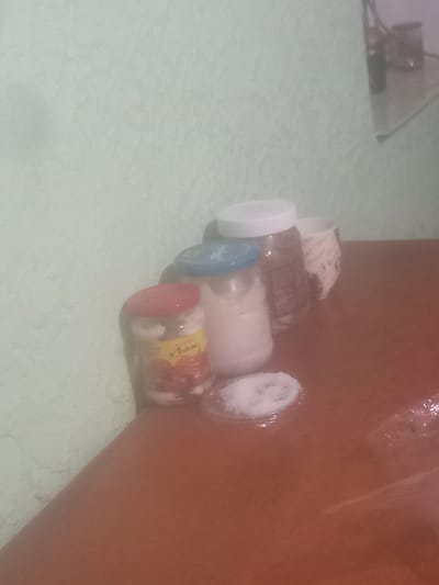 The image depicts a still life scene on a wooden surface, likely a table, against a textured, light green wall. In the foreground, a small glass jar with a red lid is filled with what appears to be preserved fruits, possibly cherries, and a white substance, possibly sugar or salt. Next to it, another glass jar with a blue lid contains a white powdery substance, likely sugar or salt, and a small dish with more of the same substance is placed in front of it. Further back, a larger jar with a white lid contains a dark substance, possibly jam or spread. To the right, a white mug with a patterned design is visible. The lighting is dim, creating shadows and a slightly hazy atmosphere. The scene is devoid of any people or explicit actions, suggesting a static arrangement of kitchen or pantry items. The location of Sîrcova, Moldova, is not visually indicated in the image itself, but the items and overall aesthetic might be consistent with a domestic setting in that region.