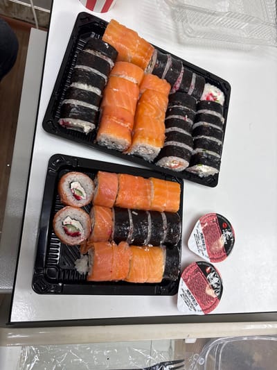 In Фролы, Russia, on a white surface, two black plastic trays of sushi are presented. The upper tray contains several rolls of sushi, some wrapped in nori seaweed and filled with rice, cream cheese, and visible red roe, while others are topped with slices of bright orange salmon. The lower tray features a similar arrangement of sushi rolls, with a focus on those topped with salmon and some with visible cream cheese and red roe. Next to the lower tray are two small, round, red-lidded containers of soy sauce or a similar condiment. The background includes a hint of a person's dark clothing and a partially visible clear plastic food container. The overall scene depicts prepared food ready for consumption.