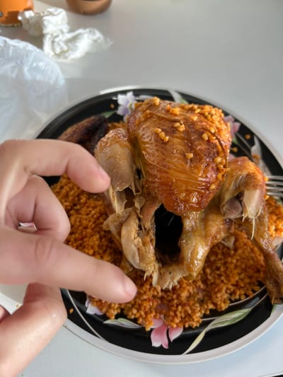The image depicts a partially consumed roasted chicken served on a bed of orange-colored bulgur or similar grain, situated on a black circular plate with a floral design. The chicken, golden-brown and exhibiting signs of internal dissection, is topped with small, light-colored颗粒, possibly nuts or spices. A left human hand, with fingers slightly curled, reaches towards the chicken, positioned centrally within the frame. A silver fork is partially visible on the right side of the plate. The plate rests on a white, unadorned surface, likely a table. In the blurred background, several domestic items are visible, including a crumpled white plastic bag, a piece of white tissue, and parts of an orange cup and a brown object. The scene occurs indoors, within the city of Illkirch-Graffenstaden, France.
