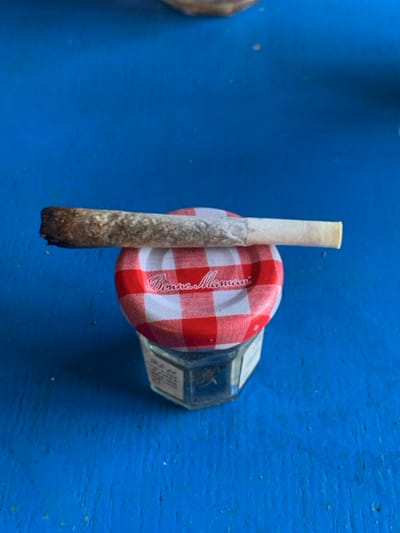 A partially smoked marijuana cigarette rests on a red and white checkered lid of a Bon Maman jam jar. The jar is octagonal and made of clear glass. The cigarette shows signs of being partially burned, with the unburnt portion a lighter color. All items are placed on a blue wooden surface.